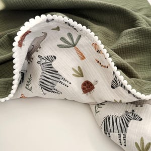 May include: A double-sided blanket featuring a solid olive green muslin fabric on one side and a white muslin fabric with a repeating pattern of zebras, turtles, and palm trees on the other. The edge is finished with a white pom-pom trim.