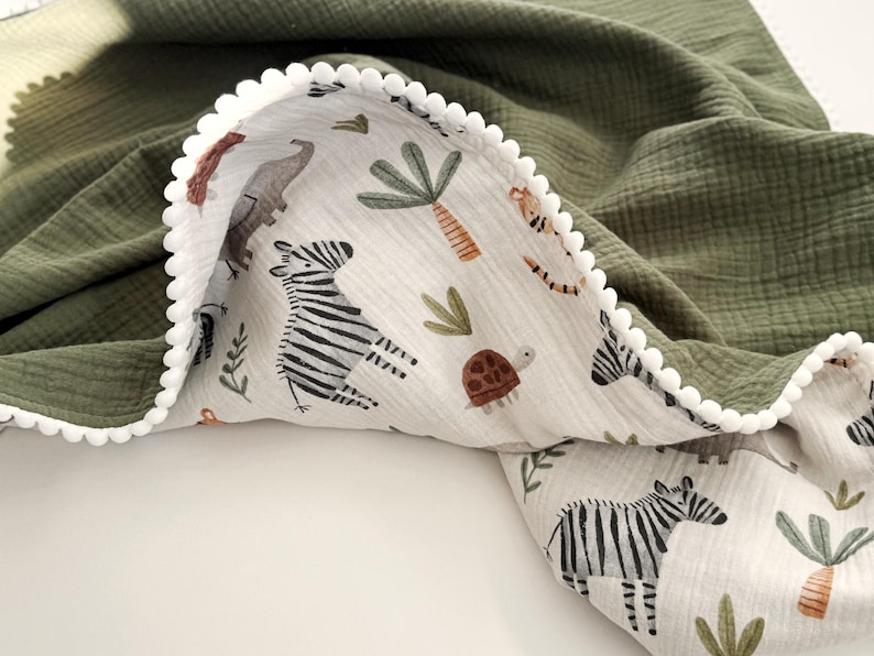 May include: A double-sided blanket featuring a solid olive green muslin fabric on one side and a white muslin fabric with a repeating pattern of zebras, turtles, and palm trees on the other. The edge is finished with a white pom-pom trim.