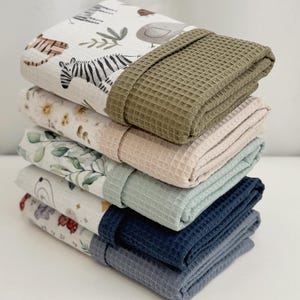 May include: A stack of five folded rectangular towels. Each towel features a patterned fabric section with animal or floral prints and a solid-colored waffle-textured fabric section in olive green, beige, sage green, navy blue, and slate blue.