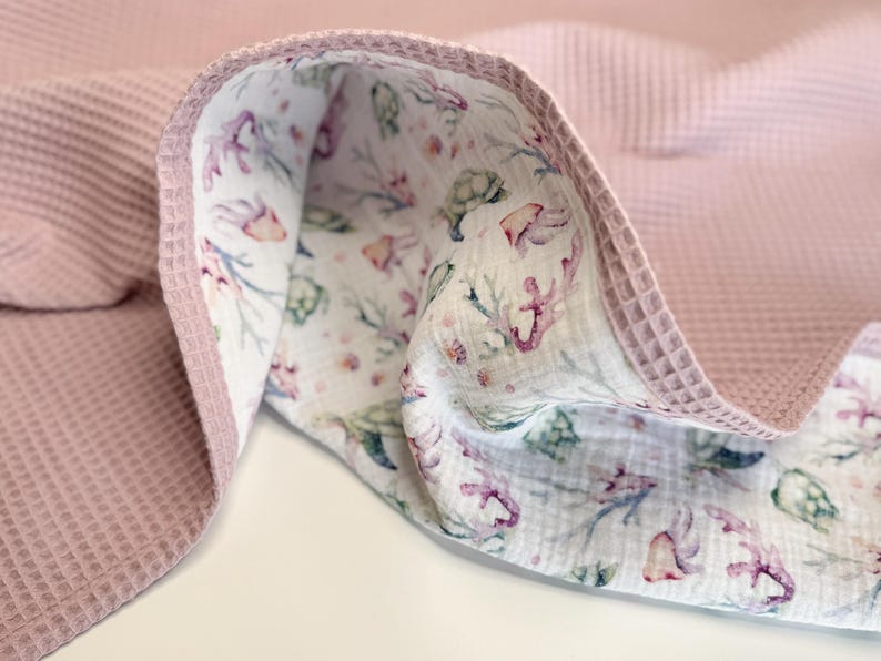 K&ouml;nnte beinhalten: Eine doppelseitige Decke mit einem altrosa Waffelstoff auf der einen Seite und einem wei&szlig;en Musselinstoff auf der anderen. Die wei&szlig;e Seite zeigt einen wiederkehrenden Aquarelldruck von Meeresschildkr&ouml;ten, Korallen und Muscheln in Rosa und Gr&uuml;n.