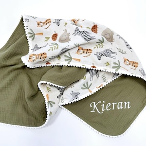 May include: A double-sided blanket featuring an olive green side with the name Kieran embroidered in white and a cream-colored side with a repeating pattern of tigers, zebras, elephants, and palm trees, finished with a white pom-pom trim.
