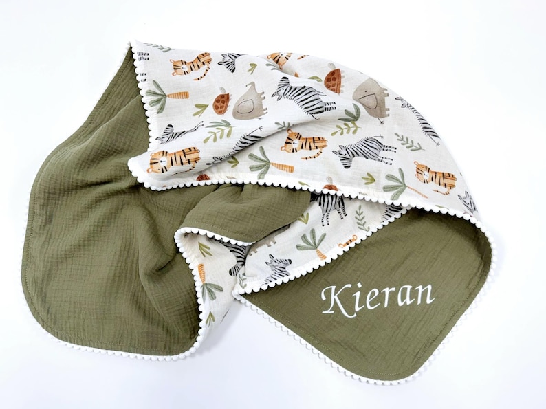 May include: A double-sided blanket featuring an olive green side with the name Kieran embroidered in white and a cream-colored side with a repeating pattern of tigers, zebras, elephants, and palm trees, finished with a white pom-pom trim.