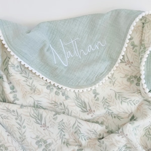 May include: A soft muslin blanket featuring a sage green solid border with white pom-pom trim and the name Nathan embroidered in white script. The main body of the blanket displays a repeating pattern of green eucalyptus leaves and small berries.