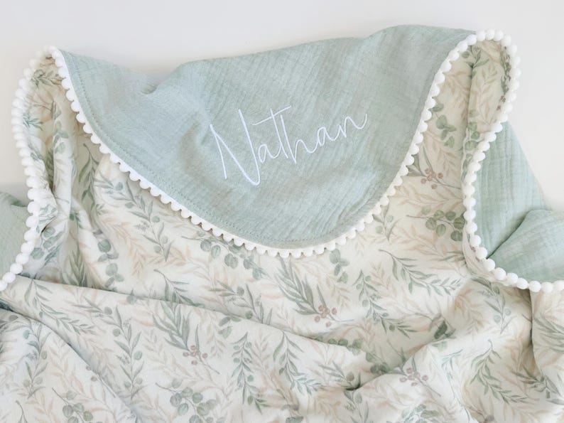 May include: A soft muslin blanket featuring a sage green solid border with white pom-pom trim and the name Nathan embroidered in white script. The main body of the blanket displays a repeating pattern of green eucalyptus leaves and small berries.