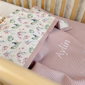 K&ouml;nnte beinhalten: Eine Babydecke aus wei&szlig;em Musselin mit einem Aquarellmuster aus gr&uuml;nen Meeresschildkr&ouml;ten und Korallen, kombiniert mit einem altrosa Waffelpiqu&eacute;-Stoff. Der Name Aylin ist in wei&szlig;er Schreibschrift auf den Waffelstoff gestickt.