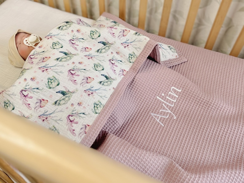 K&ouml;nnte beinhalten: Eine Babydecke aus wei&szlig;em Musselin mit einem Aquarellmuster aus gr&uuml;nen Meeresschildkr&ouml;ten und Korallen, kombiniert mit einem altrosa Waffelpiqu&eacute;-Stoff. Der Name Aylin ist in wei&szlig;er Schreibschrift auf den Waffelstoff gestickt.