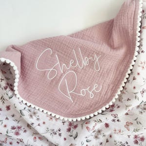May include: A soft, dusty rose-colored muslin blanket featuring the name Shelby Rose embroidered in white script. The blanket is trimmed with a white pom-pom edge and has a contrasting underside patterned with small pink flowers and butterflies.
