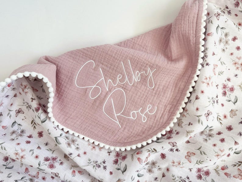May include: A soft, dusty rose-colored muslin blanket featuring the name Shelby Rose embroidered in white script. The blanket is trimmed with a white pom-pom edge and has a contrasting underside patterned with small pink flowers and butterflies.