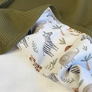 May include: A folded blanket featuring a solid olive green waffle-textured fabric on one side and a white cotton fabric on the other side, which is printed with a repeating pattern of gray elephants, black and white zebras, tigers, and palm trees.