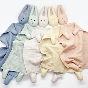 Puede incluir: Una colecci&oacute;n de mantas de seguridad para beb&eacute;s de colores pastel, con cabezas de conejito adjuntas. Las mantas son en tonos de azul, verde, rosa y amarillo. Cada conejito tiene rasgos faciales bordados. La tela parece un material ligero y texturizado.