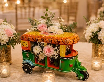 May include: A hand-painted wooden rickshaw model featuring vibrant green, orange, and yellow colors, serving as a decorative floral centerpiece filled with white chrysanthemums, pink roses, and green eucalyptus sprigs on a table.