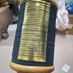 May include: A large spool of dark teal thread with a gold foil label. The spool has wooden ends and a wooden handle. The thread is tightly wound around the spool. The label has blue markings.