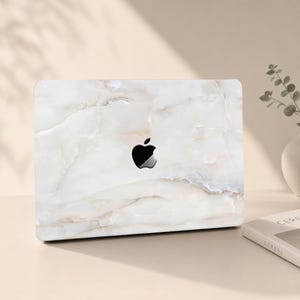 Op de afbeelding: Een laptopsticker met een wit en crèmekleurig marmerpatroon met subtiele grijze aders. Het ontwerp bedekt de buitenkant van de laptopklep, waarbij het zwarte Apple-logo in het midden zichtbaar blijft tegen de steenachtige textuur.