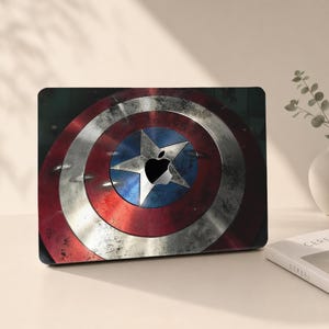 May include: A laptop skin featuring a circular shield design with a distressed red, white, and blue finish. A central white star is positioned around the cutout for the Apple logo, creating a metallic appearance with visible surface wear.