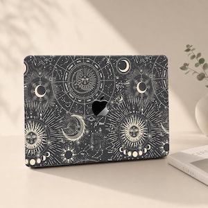 Op de afbeelding: Een zwarte harde laptophoes met een gedetailleerde witte hemelse print van zonnen, manen, sterren en een dierenriem. Het ontwerp bevat een centrale uitsparing voor het Apple-logo en toont ingewikkelde lijntekeningen van planetaire cycli.