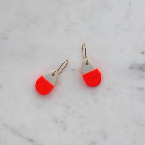Peut inclure: Une paire de petites boucles d'oreilles en forme de goutte avec un dessus blanc et un dessous orange vif. Les boucles d'oreilles sont en céramique et ont des crochets dorés.