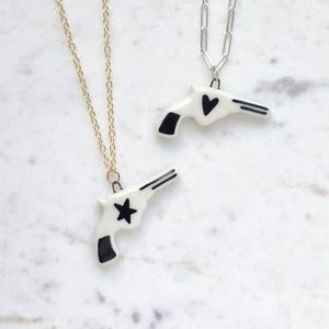 Może przedstawiać: Dwa naszyjniki z ceramicznymi zawieszkami w kształcie pistoletu. Jeden naszyjnik ma złoty łańcuszek i czarną gwiazdę na pistolecie. Drugi naszyjnik ma srebrny łańcuszek i czarne serce na pistolecie.