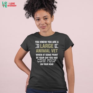 May include: A dark grey t-shirt with a white and black cow print design. The text "You know you are a large animal vet when at some point of your day you had cow poop on your head" is printed on the shirt.