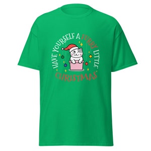 May include: A green t-shirt featuring a Christmas-themed graphic. The graphic includes a cartoon cat wearing a Santa hat, inside a pink box, with the text "Have Yourself a Furry Little Christmas" in a circular design.