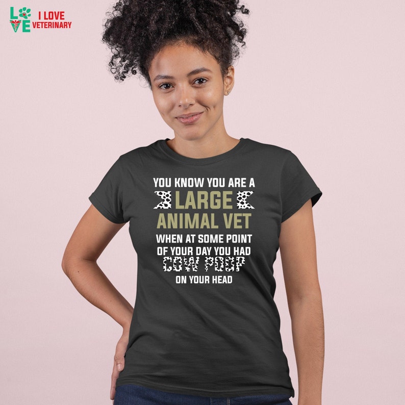 May include: A dark grey t-shirt with a white and black cow print design. The text "You know you are a large animal vet when at some point of your day you had cow poop on your head" is printed on the shirt.