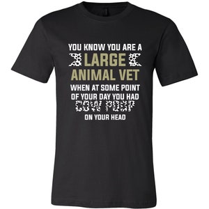 May include: Black t-shirt with a white and black cow print design. The text "You know you are a large animal vet when at some point of your day you had cow poop on your head" is printed on the shirt.