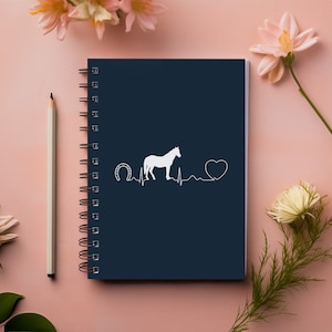 May include: A dark blue spiral notebook with a white horse silhouette, a horseshoe, and a heart beat line. The notebook is on a pink surface with flowers.