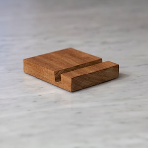 May include: A wooden phone stand with a simple design. The stand is made of light brown wood and has a rectangular shape.