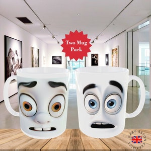 May include: Two white ceramic mugs with cartoon faces printed on them. The mugs have exaggerated facial expressions, including wide eyes, eyebrows, and mouths. The mugs are displayed on a wooden surface in front of a gallery setting. The text "Two Mug Pack" is displayed in a red starburst.