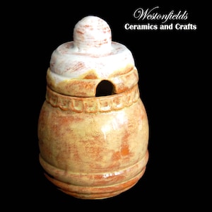 May include: A ceramic honey pot with a lid. The pot is a light brown colour with a white glaze. The lid has a white knob on top. The pot has a small hole in the lid for pouring honey.