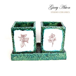 May include: Two green ceramic utensil holders with white square panels featuring brown line drawings of herbs. The holders are on a matching green tray. The text "Gary Aston STUDIO CERAMICS" is printed on a white background above the holders.
