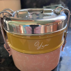 May include: A two-tiered, stainless steel and metal lunch container. The top section is mustard yellow with the word "Yum" in white script. The bottom section is pink with a white floral pattern. The container has a silver handle and clasp.