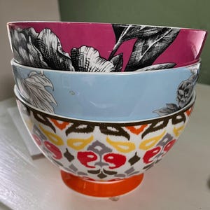 May include: A stack of three decorative ceramic bowls. The top bowl is magenta with a black and white floral design. The middle bowl is light blue. The bottom bowl has a colorful geometric pattern and an orange base.