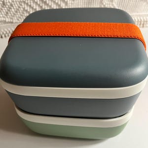 Peut inclure: Une boîte à lunch carrée empilée, avec un couvercle bleu-vert et un fond vert clair. Une bande élastique orange maintient la boîte fermée. Des bordures blanches séparent les sections.