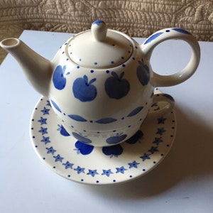 Puede incluir: Una tetera blanca con un diseño de manzanas azules y un platillo a juego. La tetera tiene una tapa y un asa. El platillo tiene un diseño de estrellas azules.