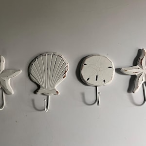 Peut inclure: Un ensemble de quatre patères murales blanches aux formes côtières, comprenant deux étoiles de mer, un coquillage et un dollar des sables. Chaque pièce présente une finition vieillie avec des bords bruns et un crochet métallique.