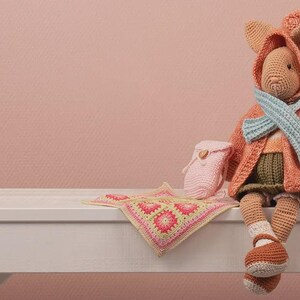 May include: A handmade crochet doll in a hooded coat, scarf, and shoes, with a small backpack and a blanket. The doll is primarily orange and tan, with a light blue scarf and green shorts. The blanket has a pink and yellow floral pattern.