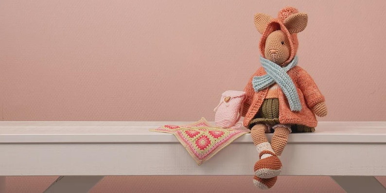 May include: A handmade crochet doll in a hooded coat, scarf, and shoes, with a small backpack and a blanket. The doll is primarily orange and tan, with a light blue scarf and green shorts. The blanket has a pink and yellow floral pattern.