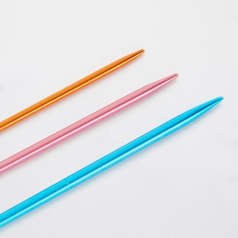 May include: Three knitting needles in different colors: orange, pink, and blue. The needles are made of metal and have a smooth, shiny finish.