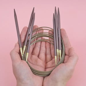 Può includere: Un set di ferri da maglia con un cavo circolare, tenuto in mani aperte su sfondo rosa. Gli aghi sono viola tenue con punte dorate. Il cavo è verde tenue abbinato. Gli aghi sono etichettati "5.0 mm/US 8".