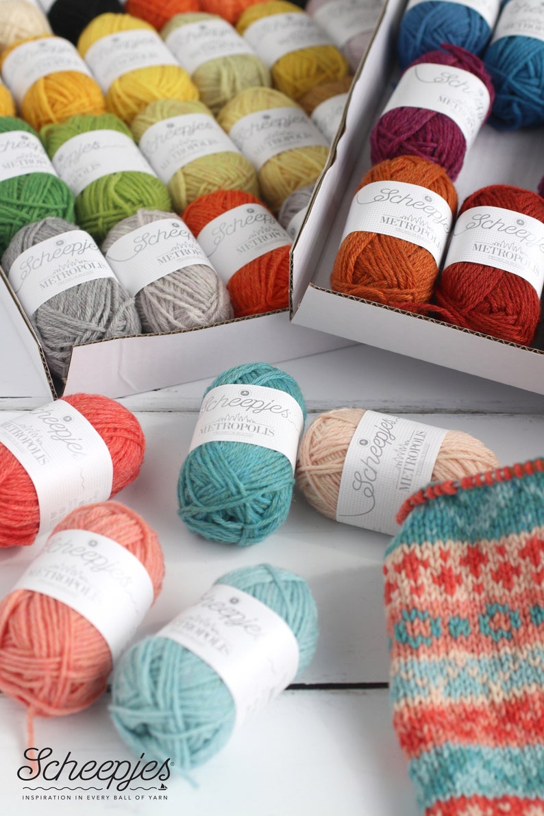 May include: A variety of colorful yarn balls in different shades of blue, green, orange, red, yellow, and pink. The yarn balls are labeled "Scheepjes Metropolis" and are arranged in a box and on a white surface. A knitted sock with a colorful pattern is also visible.