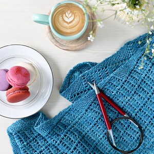 May include: A flat lay featuring a teal knitted shawl with red knitting needles, a cup of coffee with latte art, and a plate of colorful macarons. The scene is set on a white surface, with a sprig of white flowers.