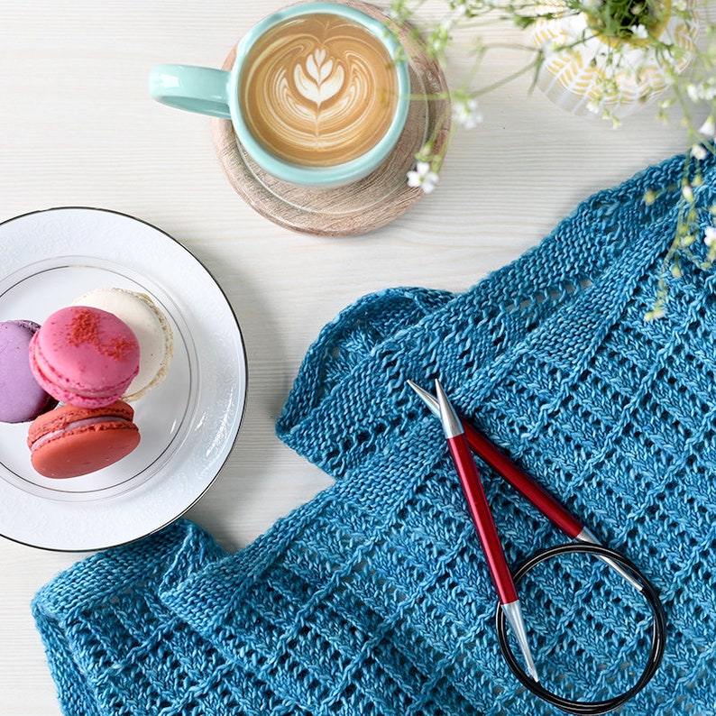 May include: A flat lay featuring a teal knitted shawl with red knitting needles, a cup of coffee with latte art, and a plate of colorful macarons. The scene is set on a white surface, with a sprig of white flowers.