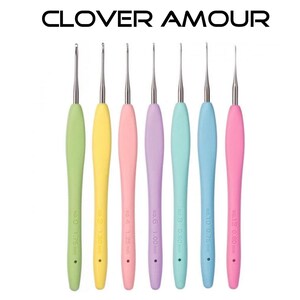Op de afbeelding: Een set van zeven haaknaalden met ergonomische handvatten in pastelkleuren: groen, geel, roze, paars, turquoise, blauw en roze. De naalden hebben zilveren metalen punten en zijn gemarkeerd met maten in millimeters. De tekst "CLOVER AMOUR" staat bovenaan.