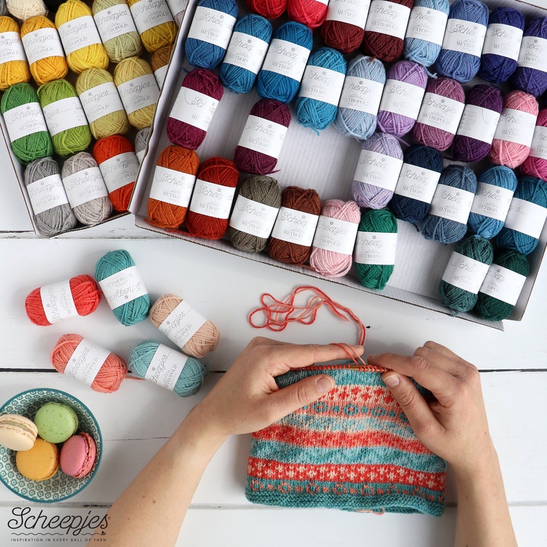 May include: A close-up of a person knitting a colorful striped hat with yarn balls in the background. The yarn balls are organized in boxes and are in various colors, including red, blue, green, yellow, and orange. The yarn is labeled "Scheepjes".