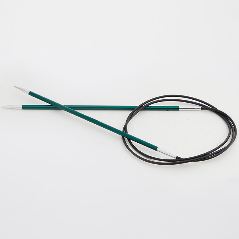 May include: A pair of green circular knitting needles with silver tips and a black flexible cable. The needles are lying on a white background. The needles are a crafting tool.