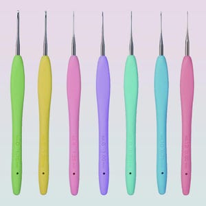 Op de afbeelding: Een set van acht haaknaalden met gekleurde handvatten in groen, geel, roze, paars en blauw. Elke haaknaald heeft een zilveren metalen punt en is gemarkeerd met een maat in millimeters, variërend van 0,60 mm tot 1,75 mm.