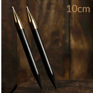 May include: A pair of black knitting needles with gold-coloured tips. The needles are 10 cm (3.9 inches) long. The needles are angled towards each other, set against a dark wooden background.