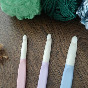 May include: Three crochet hooks with white heads and pastel-colored handles in pink, lavender, and light blue. The hooks are arranged vertically, each featuring a smooth, ergonomic design with a distinct hook tip for yarn manipulation.