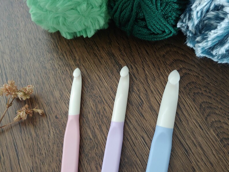 May include: Three crochet hooks with white heads and pastel-colored handles in pink, lavender, and light blue. The hooks are arranged vertically, each featuring a smooth, ergonomic design with a distinct hook tip for yarn manipulation.