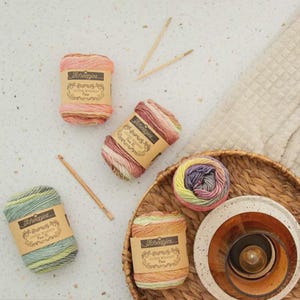 May include: Four skeins of Scheepjes Stone Washed yarn in variegated pastel colorways, each with a paper label. Included are a wooden crochet hook and a pair of circular knitting needles. One skein is unwound, resting in a woven wicker basket.